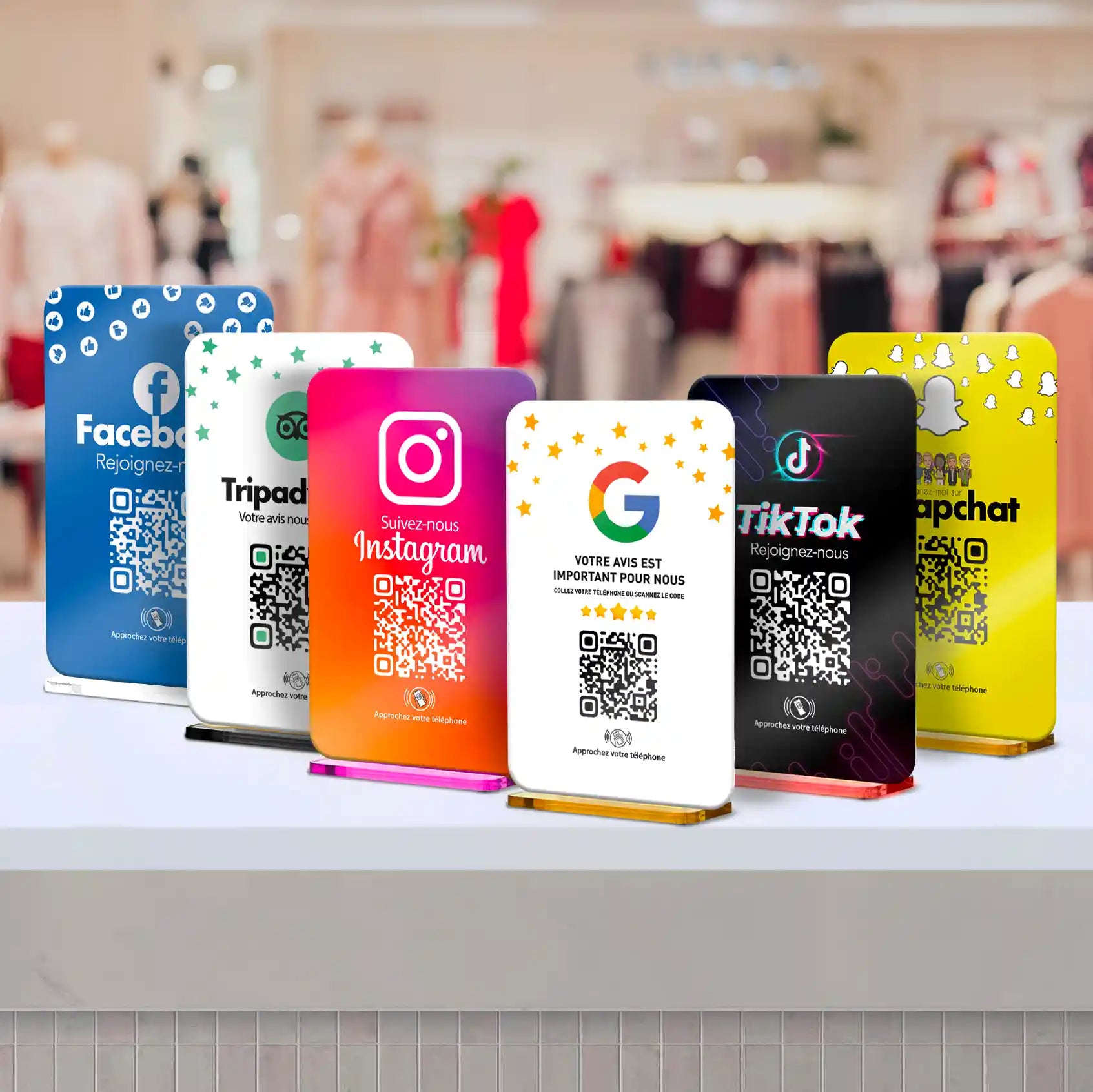 Plaques NFC et QR Code colorées avec logos de réseaux sociaux tels que Facebook, Instagram, TikTok, Snapchat, Google et Tripadvisor, sur un comptoir blanc dans un environnement flou de boutique.