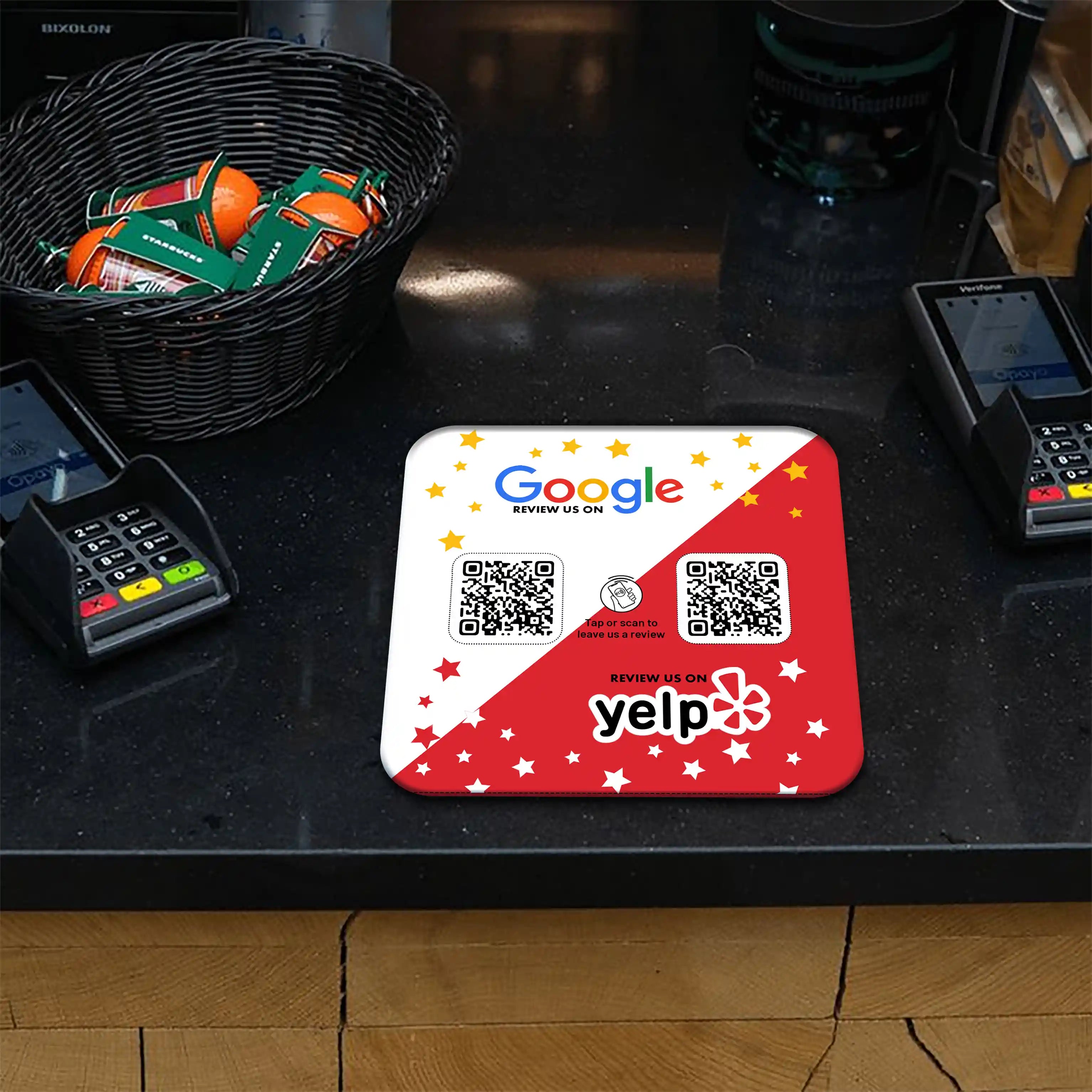 Plaque avec des codes QR pour les avis sur Google et Yelp, posée sur un comptoir noir à côté de terminaux de paiement. Panier en osier contenant des oranges avec des petites étiquettes vertes. Ambiance de comptoir professionnel.