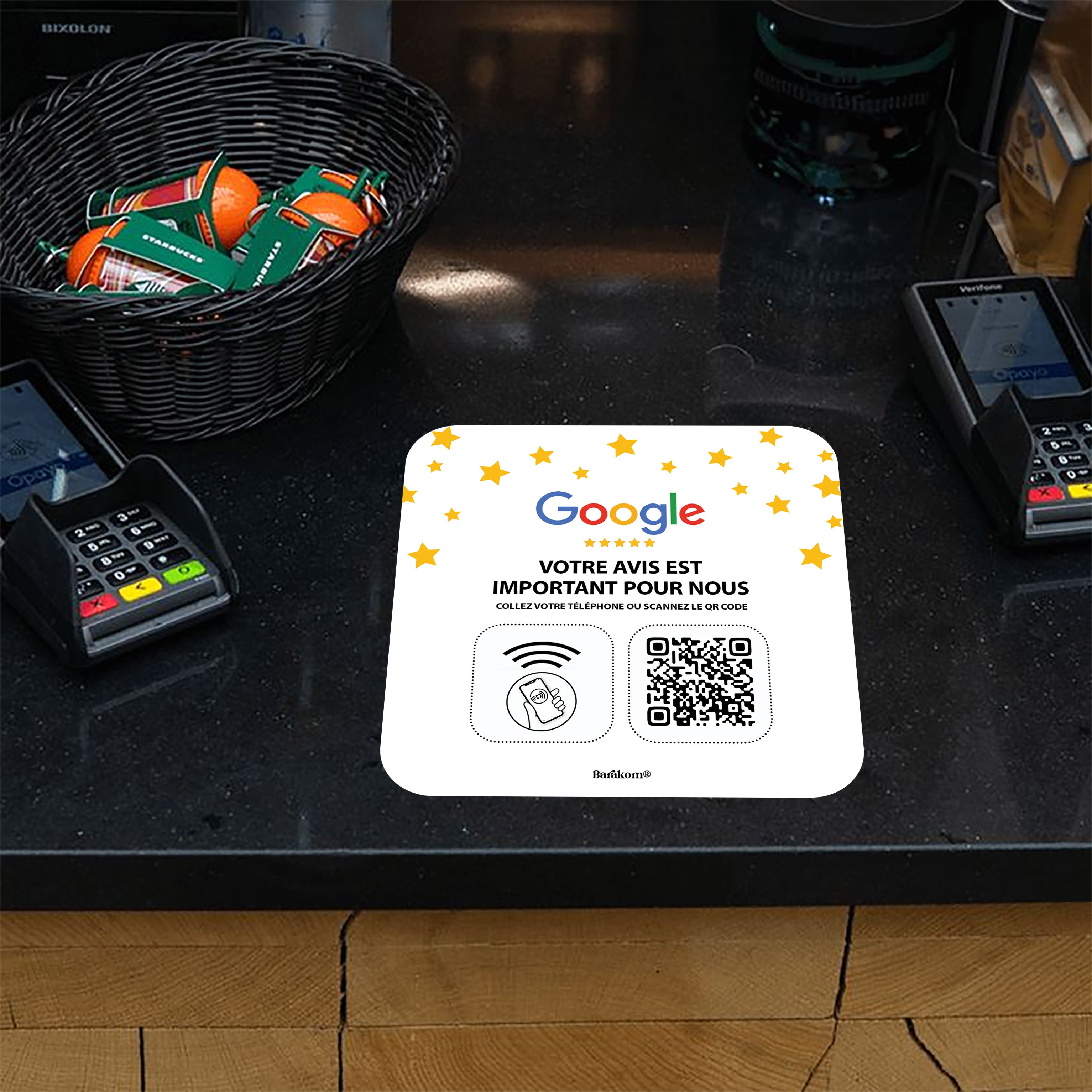 Plaque avec NFC et QR Code posée sur un comptoir, entourée de terminaux de paiement et d'un panier de friandises, encourageant les avis Google pour Barakom.
