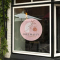 Vitrine de magasin avec autocollant de signalétique circulaire, personnalisée pour "Emily Beauty Cosmetics". Design minimaliste avec illustration florale sur fond rose pâle, positionnée sur une grande fenêtre en verre. Idéal pour les commerces et artisans cherchant une vitrine attrayante et moderne.