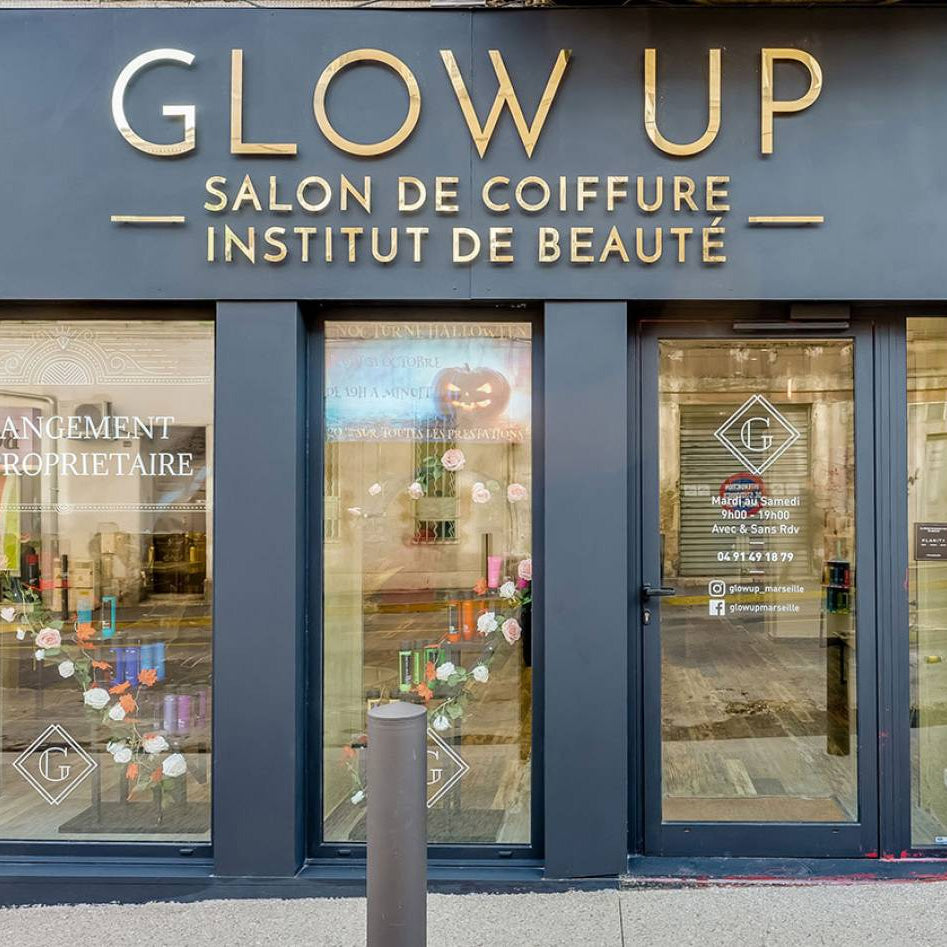 Vitrine de salon de coiffure 'Glow Up' avec signalétique dorée, décorée de fleurs et produits cosmétiques visibles à travers les vitres. Le salon propose également des services d'institut de beauté.