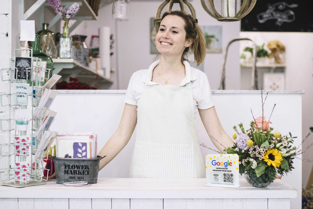 Fleuriste souriante tenant une plaque NFC et QR code sur le comptoir avec un bouquet de fleurs à côté, dans un magasin de fleurs. Plaques NFC et QR code pour avis clients. Décoration intérieure florale.