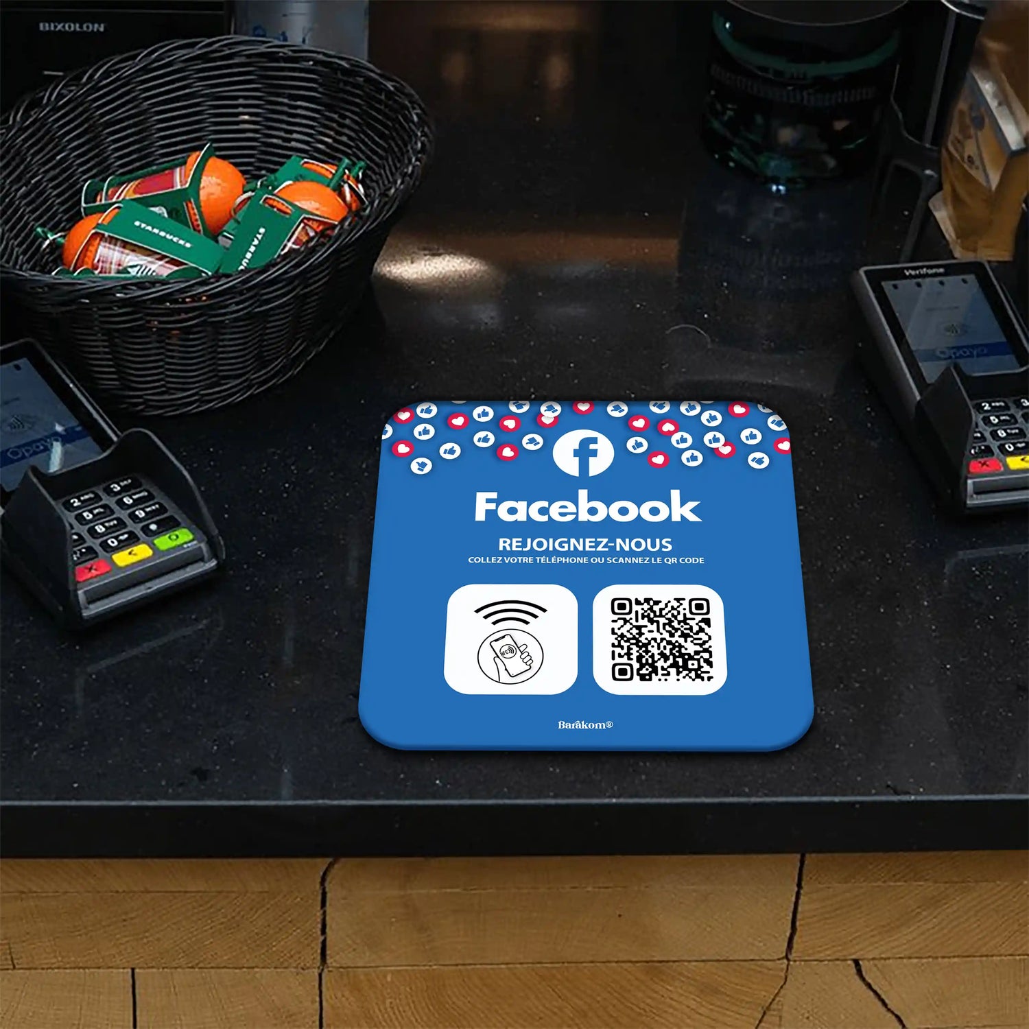 Plaque NFC et QR Code sur le comptoir d'un magasin avec terminal de paiement, incitant à rejoindre une page Facebook en scannant le code ou en utilisant NFC, entourée d'un panier de bonbons.