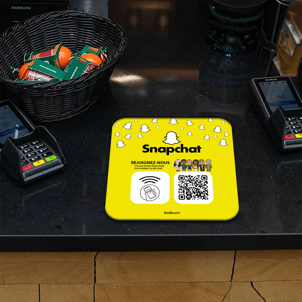 Plaque NFC et QR Code jaune Snapchat sur un comptoir, entourée de terminaux de paiement. Design invitant à rejoindre via contact téléphonique ou scan du code, accompagné de dessins animés et logo Snapchat. Panier de fruits en arrière-plan.