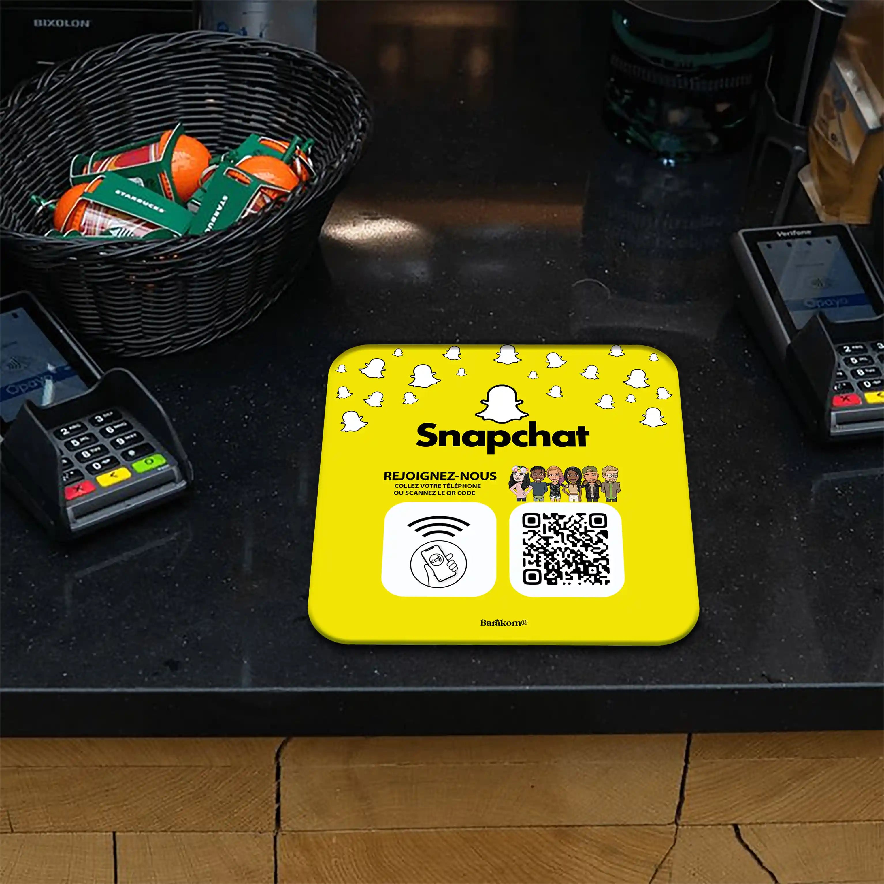 Plaque NFC et QR Code jaune Snapchat sur un comptoir, entourée de terminaux de paiement. Design invitant à rejoindre via contact téléphonique ou scan du code, accompagné de dessins animés et logo Snapchat. Panier de fruits en arrière-plan.