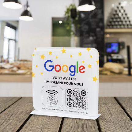 Plaque interactive NFC et QR Code sur une table en bois dans un café moderne, avec des lumières pendantes et un comptoir en arrière-plan. Texte sur la plaque : 'Votre avis est important pour nous'.",