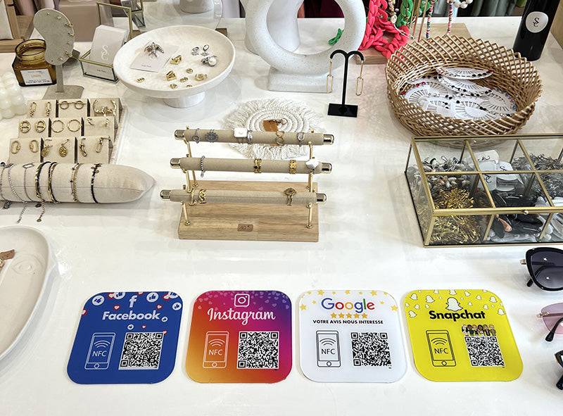 Plaques NFC et QR codes adhésives pour réseaux sociaux Facebook, Instagram, Google et Snapchat, disposées sur une table avec bijoux en exposition.